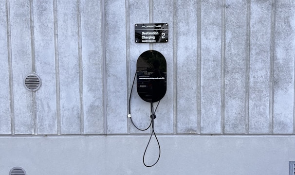 A modern electric vehicle charging station is mounted on a vertical concrete wall. The station is sleek and black, with a connected charging cable hanging down. The concrete wall has vertical grooves and is slightly weathered. The ground is paved with cobblestones, and there are small circular vents on the wall.
