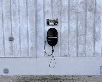 A modern electric vehicle charging station is mounted on a vertical concrete wall. The station is sleek and black, with a connected charging cable hanging down. The concrete wall has vertical grooves and is slightly weathered. The ground is paved with cobblestones, and there are small circular vents on the wall.