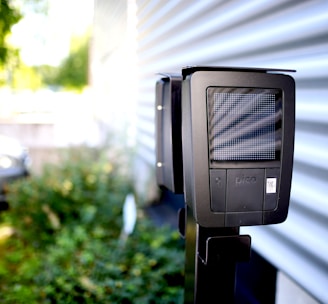 A dark-colored charging station with a digital display is installed outdoors next to a corrugated metal wall. In the background, a white car is partially visible in a parking area surrounded by greenery.