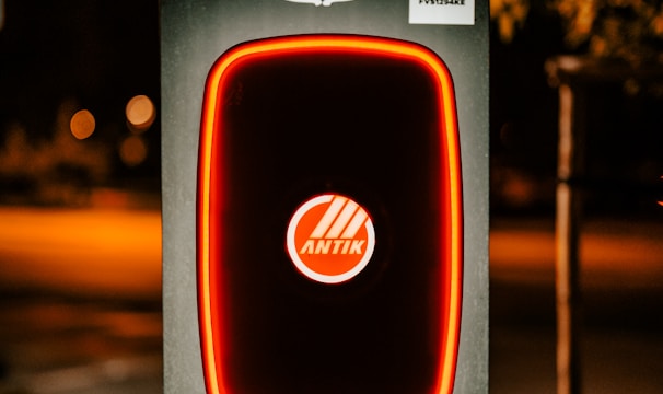 A modern electric vehicle charging station is illuminated at night. The charger has bold red lighting outlining its frame. Above the charger, there is a detailed and colorful coat of arms. The background is dimly lit, suggesting it's a suburban area or a park.