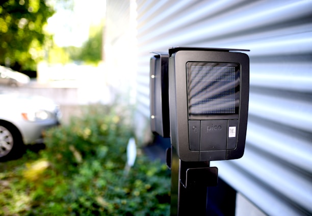 A dark-colored charging station with a digital display is installed outdoors next to a corrugated metal wall. In the background, a white car is partially visible in a parking area surrounded by greenery.