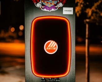 A modern electric vehicle charging station is illuminated at night. The charger has bold red lighting outlining its frame. Above the charger, there is a detailed and colorful coat of arms. The background is dimly lit, suggesting it's a suburban area or a park.